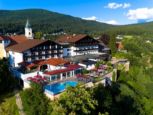 Wir sind derzeit auf der Suche nach Verstärkung für unser Rezeptionsteam Außenansicht des Wellnesshotels Hofbräuhaus in Bodenmais – Panoramablick auf Hotel, Außenpool, Saunen und Bayerischen Wald bei Sonnenschein