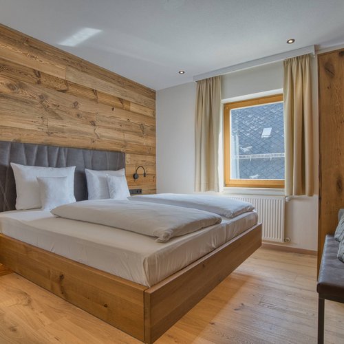 Häufige Fragen zum Wellnesshotel Hofbräuhaus in Bodenmais Helles zweites Schlafzimmer einer Suite im Wellnesshotel Hofbräuhaus in Bodenmais, ausgestattet mit komfortablem Doppelbett, Holzverkleidung, Fenster mit Blick auf das Dach und die umliegende Natur