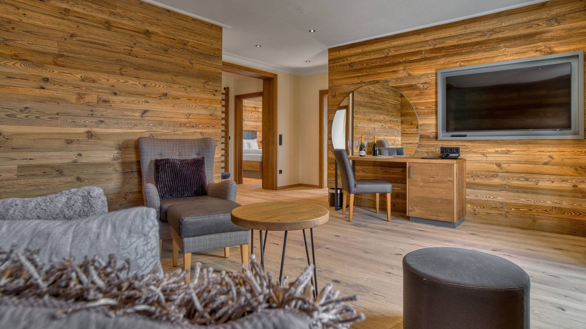 Ihr Traumurlaub im Hotel Hofbräuhaus Gemütlicher Wohnbereich einer Suite im Wellnesshotel Hofbräuhaus in Bodenmais mit moderner Couch, Sessel, Flachbild-TV, großem runden Spiegel.